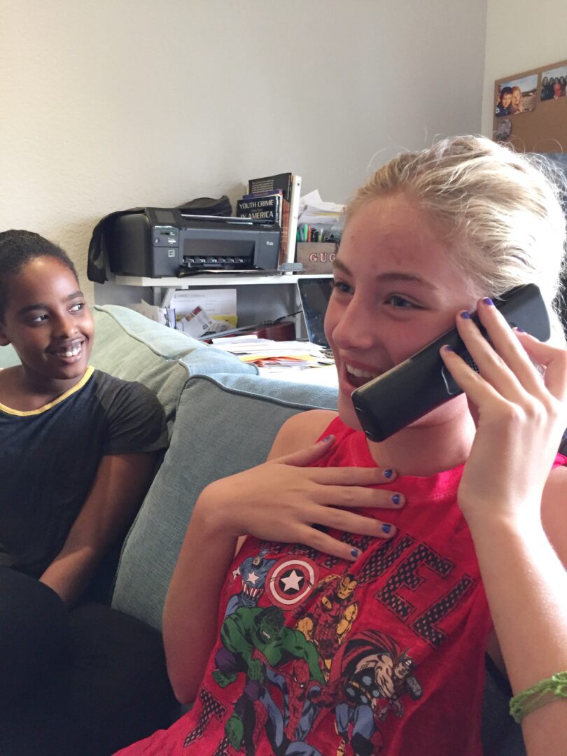 Two girls sitting on a couch talking on the phone.