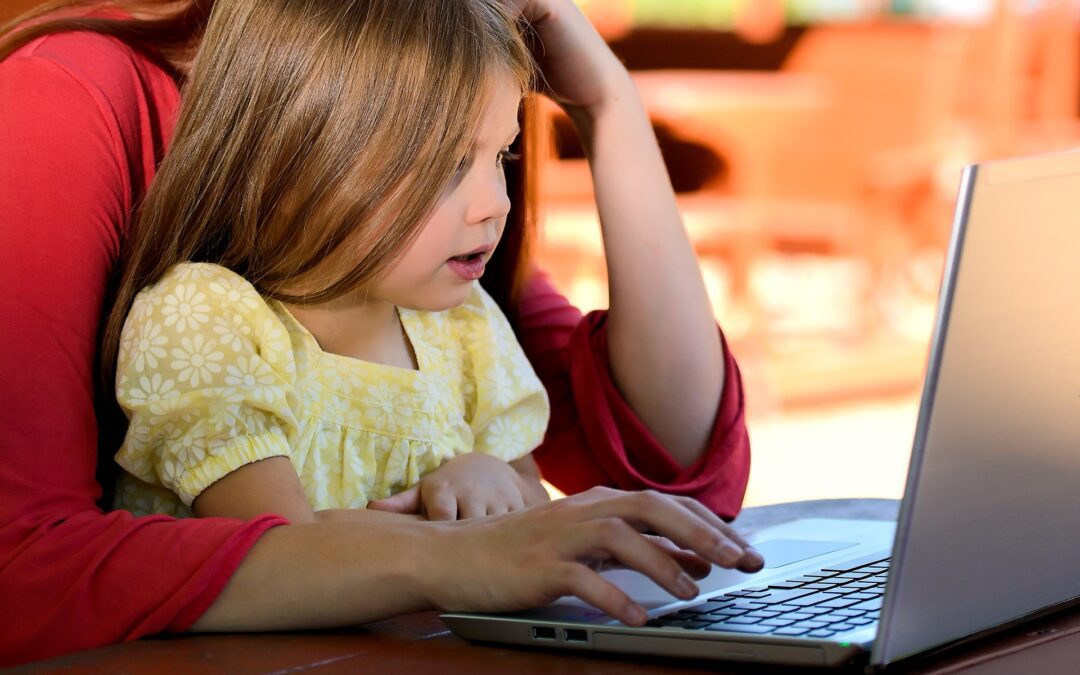 A little girl sitting at the table with her mother on a laptop.